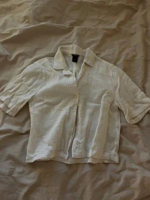 Vit kortärmad skjorta från Lindex - Säljer en vit, luftig skjorta från Lindex med korta ärmar och knappar framtill. Skjortan har en krage och är tillverkad i mjukt, strukturerat bomullstyg som ger en chill och avslappnad vibe. Passformen är loose och skjortan är något croppad.