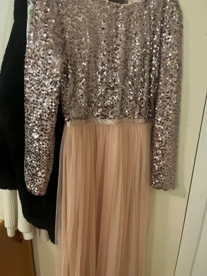 Långklänning med paljetter och tyll - Superfin långklänning i ljus champagnefärg med glittrig paljettöverdel och lång, flowy kjol i tyll. Klänningen har långa ärmar och en elegant, skimrande look som verkligen sticker ut. Perfekt för dig som vill känna dig extra glam.