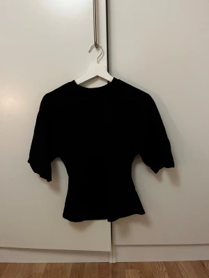 Svart topp från Lager 157, figursydd XS - Svart topp från Lager 157 i figursydd modell med korta ärmar och rund halsringning. Enkel och stilren design som sitter snyggt i midjan. Perfekt att matcha med jeans eller kjol för en clean look.