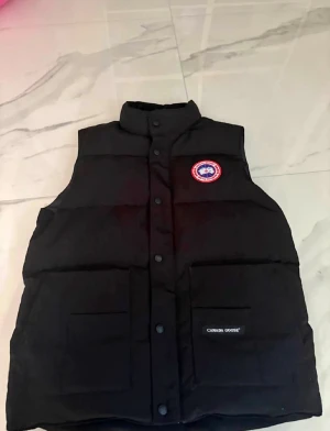 Svart dunväst från Canada Goose - Svart dunväst från Canada Goose med hög krage och tryckknappar framtill. Västen har två stora fickor på framsidan, varav en med Canada Goose-logga. Klassisk rund logotyp på bröstet. Perfekt för kyliga dagar och enkel att styla över en hoodie. ÄKTA KOM MED BUD! Dm för flera bilder/videos