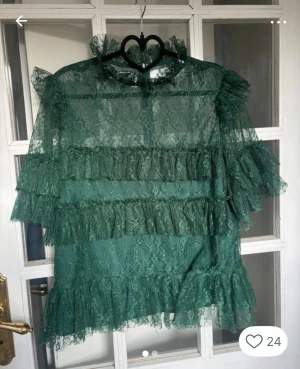 Grön spetsblus med volanger By Malina - Supersnygg grön spetsblus från By Malina med volanger och hög krage. Blusen är genombruten med flera lager volangdetaljer och har korta ärmar. Perfekt för dig som vill sticka ut med en romantisk och trendig look.