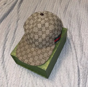 Gucci keps - Säljer en beige Gucci keps med klassiskt GG-mönster och grönt/rött band på sidan. Kepsen är tillverkad i bomull och har en snygg, rundad form med böjd skärm. Perfekt för dig som vill ha en lyxig och trendig accessoar. Pris kan däremot diskuteras.