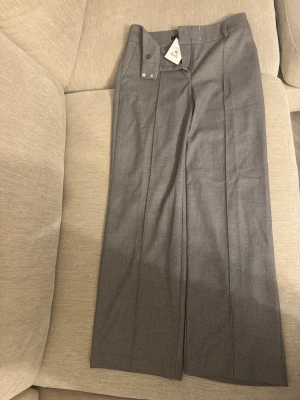 Grå kostymbyxor från Bik Bok, strl 38 - Snygga grå kostymbyxor från Bik Bok i storlek 38. Byxorna har raka ben, pressveck och klassisk midja med hällor för bälte. Materialet är mjukt och har en stilren finish, perfekt för en clean och modern look.