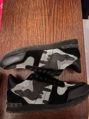 Valentino Garavani rockrunners  - Snygga svarta och vita sneakers från Valentino Garavani med coolt camouflage-mönster. Skorna har snörning, platt sula och detaljer i mocka och syntet. Perfekt för dig som vill sticka ut med en unik design och exklusivt märke.(pris kan diskuteras om det är en snabb affär)