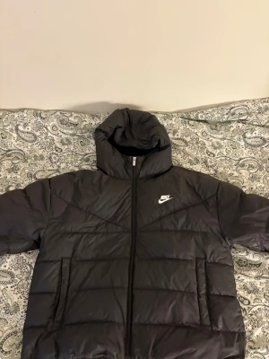 Svart pufferjacka från Nike Storm-FIT - Svart pufferjacka från Nike Storm fit, huva och vit Nike-logga på bröstet. Jackan har dragkedja framtill, två sidofickor och innerficka. Köpt 2022 för 2500kr, använd en vinter och fåtal gånger efter det:)
