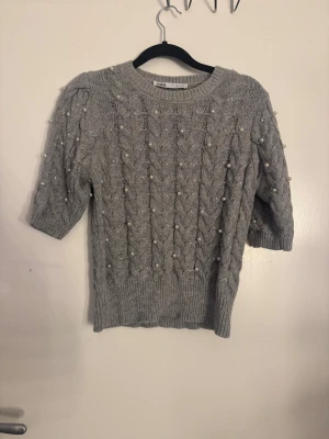 Grå stickad tröja med pärlor från Zara - Supersöt grå stickad tröja från Zara med korta ärmar och snyggt flätmönster. Tröjan är dekorerad med pärlor över hela framsidan och ärmarna, vilket ger en extra touch. Rund hals och ribbade muddar. Perfekt för dig som gillar detaljer och vill ha något unikt i garderoben.