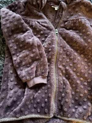 Plommonlila fleecejacka med ”prickar” från Pompdelux - Supermjuk fleecejacka i plommonlila från Pompdelux, storlek 128. Jackan har ett prickigt mönster, gosig krage och glittriga kanter. Dragkedja framtill och ribbade muddar vid ärmslut. Perfekt för dig som gillar mysiga och coola detaljer.