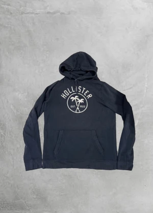 Mörkblå Hollister hoodie med tryck - Snygg mörkblå hoodie från Hollister med stort vitt tryck på bröstet med palmträd och texten 'Hollister Est. 1922'. Tröjan har huva med snörning och en klassisk magficka. Perfekt för chill dagar och enkel att matcha med jeans eller joggers.