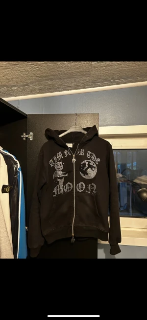 Svart hoodie Aim For The Moon - Svart hoodie med tryck i silver på bröstet och coolt motiv. Dragkedja framtill med unik zip-puller och två fickor. Huva och ribbade muddar. Perfekt för dig som gillar streetwear och vill sticka ut.