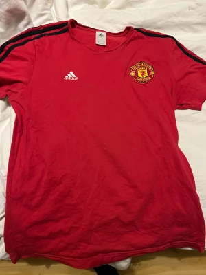Manchester United röd Adidas fotbollströja - Röd Manchester United fotbollströja från Adidas med klubbmärke på bröstet och vita Adidas-loggan. Svarta ränder längs axlarna ger en sportig vibe. Tröjan är kortärmad och har rund hals. Perfekt för dig som vill representera United både på match och träning. Är 185 cm lång o väger 80kg