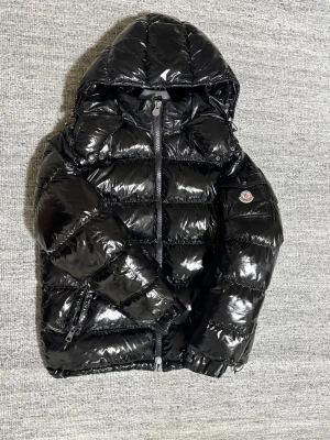 Svart Moncler pufferjacka med huva - Säljer en svart, glansig pufferjacka från Moncler med huva och dragkedja framtill. Jackan har Moncler-logga på ärmen, quiltad design och innerfoder med serietidningsmönster. Perfekt för dig som vill ha en varm och snygg vinterjacka med street vibe.