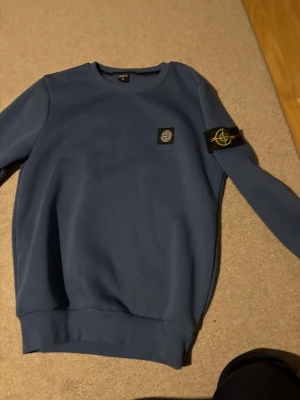 Blå Stone Island sweatshirt - Säljer en blå crewneck sweatshirt från Stone Island med klassisk patch på vänster ärm och logga på bröstet. Tröjan har rund hals, ribbade muddar och är tillverkad i mjuk bomull. Perfekt för dig som gillar streetwear och vill ha något stilrent.pris diskuteras