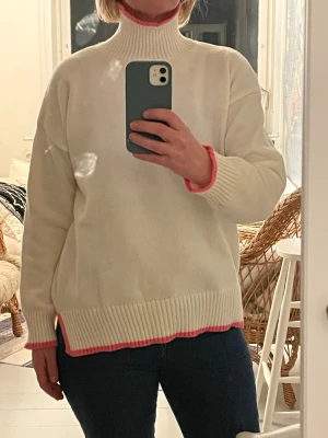 Vit polotröja med rosa kant Meet Me There - Säljer en off white polotröja från Meet Me There med ribbad hög krage och snygga rosa detaljer vid hals, ärmslut och nederkant. Tröjan är stickad och har långärmad passform med slits på sidorna. Köpte direkt från leverantören, aldrig använd. 