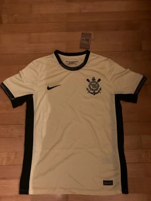 Gul och svart Corinthians fotbollströja Nike - Snygg Corinthians fotbollströja från Nike i gult med svarta detaljer längs sidorna, ärmslut och krage. Tröjan har klubbens emblem på bröstet och Nike-logga. Tillverkad i lätt och ventilerande polyester, perfekt för match eller träning.