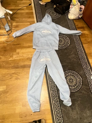 Cortiez ljusblå tracksuit  - Ljusblå hoodie med huva och magficka, samt ett vitt tryck med motiv av ett fartyg på bröstet. Tröjan har långa ärmar och är gjord i mjukt material, perfekt för chill dagar. Snygg och enkel design som passar till jeans eller mjukisbyxor.