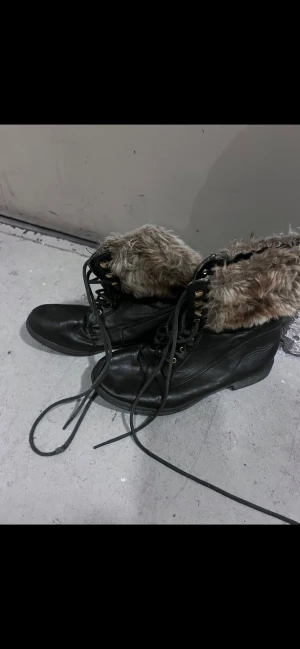Svarta boots med fuskpäls och snörning - Säljer ett par svarta boots med snörning och detaljer i guld. Insidan och kanten är fodrad med mjuk, beige fuskpäls som ger extra värme och cool stil. Skorna har platt sula och rund tå, perfekta för kalla dagar. Materialet är skinnimitation och päls.