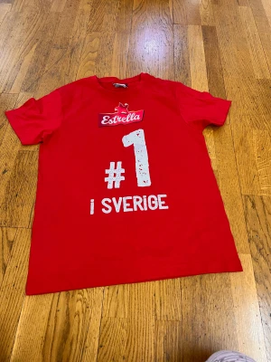 Röd Estrella t-shirt från Clique - Säljer en röd t-shirt från Clique i storlek S med tryck 'Estrella #1 i Sverige' på framsidan. T-shirten har rund hals och korta ärmar, tillverkad i mjuk bomull. Perfekt för dig som gillar snacksvibes och vill sticka ut med en cool printad front.