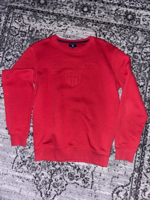 Röd sweatshirt från GANT - Snygg röd sweatshirt från GANT med broderad logga och emblem på bröstet. Tröjan har rund hals, ribbade muddar och är tillverkad i mjuk bomull. Perfekt för dig som gillar klassisk stil med en modern touch.