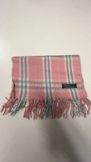 Rutig rosa halsduk från Burberrys - Säljer en klassisk rutig halsduk från Burberrys i mjuk lammull. Halsduken är rosa med ljusblå och beige rutor samt fransar i ändarna. Perfekt accessoar för att lyfta din outfit med en touch av brittisk stil. En liten defekt på halsduken som kan ses på bilden!