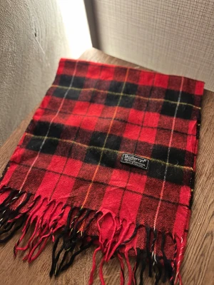 Rutig röd halsduk från Burberrys - Klassisk rutig halsduk från Burberrys i 100% ull. Halsduken har fransar i ändarna och den ikoniska Burberrys-etiketten. Perfekt accessoar för att lyfta din outfit. Anledningen att de stör ”burberrys” är för att de är en äldre modell.