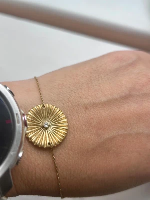 Edblad armband med solformad berlock - Snyggt armband i stål guldton med tunn kedja och en rund, solformad berlock med räfflad yta. I mitten sitter en liten klar sten som ger extra bling. Perfekt för dig som gillar unika detaljer och stilren design.