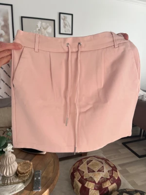 Ljusrosa kortkjol från ONLY - Snygg ljusrosa kortkjol från ONLY med snörning i midjan och breda hällor. Kjolen har en enkel, rak form och är gjord i mjukt bomullsmaterial som känns skönt mot huden. Perfekt för dig som gillar en clean och chill stil.