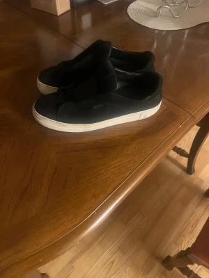 Svarta sneakers från Axel Arigato - Svarta sneakers från Axel Arigato med stilren design och bred elastisk rem över foten. Skorna har en vit platt sula och är tillverkade i mocka med diskret logga på sidan. Perfekt för dig som gillar minimalistisk och modern stil.