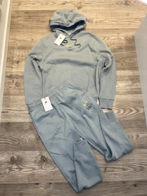 Nike tracksuit - Nike tracksuit med stor gul Swoosh och svart text på ryggen. Framtill finns mindre tryck och logga. Tröjan har huva med snörning, ribbade muddar och är tillverkad i mjuk bomullsmix. Perfekt för chill eller streetwear. Kvitto finns