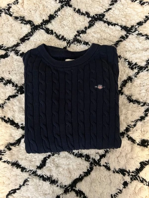Mörkblå kabelstickad tröja från GANT - Säljer en mörkblå kabelstickad tröja från GANT med rund hals och broderad logga på bröstet. Tröjan är i mjuk bomull och har långa ärmar samt ribbade muddar. Perfekt för dig som gillar klassisk och stilren look.