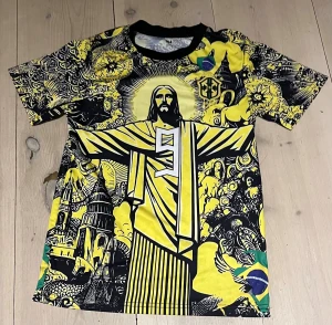 Ronaldo #9 Brasil grafisk fotbollströja - Unik fotbollströja med Ronaldo #9 och Brasilien-tema. Färgstark design i gult, svart och inslag av grönt och blått, med stora grafiska tryck av Kristusstatyn och brasilianska symboler. Korta ärmar och rund hals. Perfekt för dig som vill sticka ut på match eller i stan.