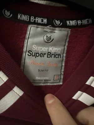 Vinröd sweatshirt King B-Rich S - Snygg vinröd sweatshirt från King B-Rich i slim fit. Tröjan har vita grafiska detaljer framtill och en svart triangel med romerska siffror på ryggen. Tillverkad i mjuk bomull med långa ärmar och ribbade muddar. Perfekt för dig som gillar streetwear och unik design.