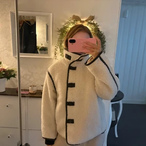 Vit teddyjacka med svarta detaljer - Säljer en fluffig vit teddyjacka med svarta kantband och coola knäppningar framtill. Jackan har hög krage och långärmade ärmar, perfekt för att hålla sig varm och se stylish ut. Passformen är loose och jackan har en unik look som sticker ut.