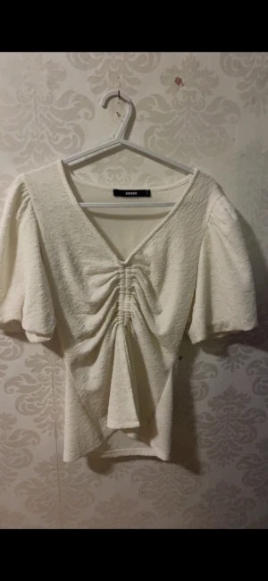 Vit omlottopp med puffärm från Bik Bok - Supersöt vit omlottopp från Bik Bok med puffiga korta ärmar och snygg snörning framtill som ger en cool rynkad effekt. Materialet är mjukt och har en lite strukturerad yta. V-ringad och figurnära passform, perfekt till jeans eller kjol. Har dock en liten fläck men går till 97% chans bort med fläckborttagning. Men den syns knappt 