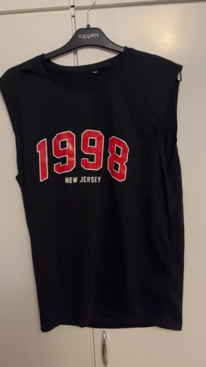 Svart ärmlös t-shirt 1998 New Jersey - Svart ärmlös t-shirt från KappAhl med tryck i rött och vitt där det står '1998 New Jersey' på bröstet. Klassisk rund halsringning och rak passform. Perfekt för dig som gillar sportig och retro stil.