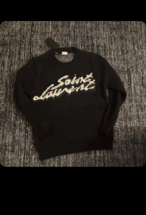 Svart Saint Laurent stickad tröja - Svart stickad tröja från Saint Laurent med vit logotyp framtill i en cool, handskriven stil. Tröjan har rund hals och långa ärmar, perfekt för dig som gillar exklusiva märken och vill ha en unik look. Mjuk och fluffig känsla som gör den extra skön. Helt ny testad bara 