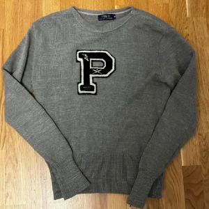 Ralph Lauren Tröja - Grå stickad tröja från Polo Ralph Lauren med svartvit P-applikation. Klassisk college-stil med rund hals och ribbade muddar. Midjan har krympt något – perfekt för slim fit och passar även Small.