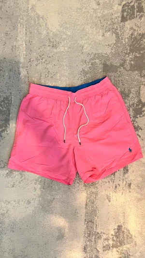 Rosa badshorts från Polo Ralph Lauren - Snygga rosa badshorts från Polo Ralph Lauren med vit snörning i midjan och klassisk blå logga broderad på benet. Shortsen har elastisk midja, innerbyxa och en bakficka med knapp. Perfekta för stranden eller poolen.