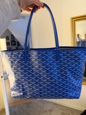 Blå Goyard väska  - Säljer en ikonisk blå shoppingväska från Goyard med deras klassiska grafiska mönster i vitt. Väskan har långa handtag, rymlig insida och är tillverkad i slitstarkt syntetmaterial med skinn-detaljer. Perfekt för dig som vill sticka ut med en lyxig och trendig accessoar. Ingår en clutch som är super snygg även att ha separat! Det är en AA-kopia!