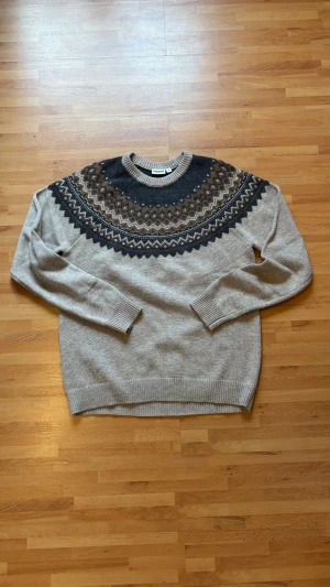 Beige stickad tröja med mönster - Snygg stickad tröja från Kappahl i beige med mörkgrått och brunt mönster runt halsen. Rund halsringning och långa ärmar med ribbade muddar. Perfekt för dig som gillar klassisk nordisk stil och vill ha något varmt och skönt.