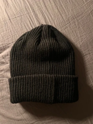 Svart ribbstickad mössa Canada Goose (Ganska liten storlek!) - Svart ribbstickad mössa från Canada Goose med klassisk rund patch framtill i rött, vitt och blått. Mössan har uppvikt kant och är gjord i mjukt, värmande material som passar perfekt när det är kallt ute. Snygg och enkel design med streetkänsla.