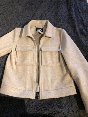 Beige kappa/overshirt  från Pieces - Säljer en stilren beige overshirt från Pieces med klassisk krage, två stora fickor framtill och dragkedja. Jackan har lång ärm och en rak, normal passform. Perfekt att slänga på över en hoodie eller t-shirt för en chill look.