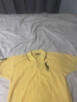 Gul pike från Ralph Lauren - Säljer en gul polotopp från Ralph Lauren med svart polospelare-logga på bröstet och rygg samt siffran 3 på ärmen. Toppens krage har svarta detaljer och tre knappar framtill. Materialet är mjuk bomull och modellen är klassisk med korta ärmar.