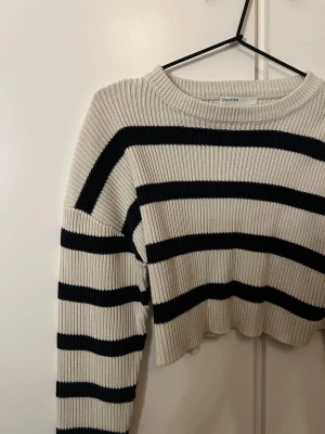 Randig croppad stickad tröja Bershka - Superfin croppad stickad tröja från Bershka i beige och svart med breda ränder. Tröjan har ribbad struktur, rund hals och långa ärmar. Perfekt för dig som gillar en chill och trendig look.