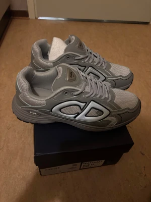 Grå Dior sneakers med CD-detalj - Snygga grå sneakers från Dior med stora CD-detaljer på sidorna. Skorna har meshpaneler och partier i syntetmaterial, samt snörning framtill. Sulan är chunky och ger en modern vibe. Perfekta för dig som vill sticka ut med exklusiva och trendiga sneakers.