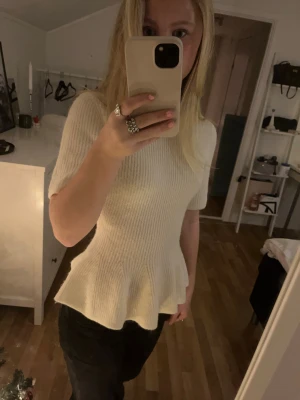 Vit stickad topp med volangkant - Säljer en superfin vit stickad topp med korta ärmar och volangkant nedtill. Toppen har en ribbad struktur och sitter snyggt i midjan för en feminin siluett. Perfekt att styla med jeans eller kjol för en trendig look.