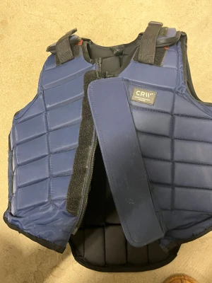 Blå CRW skyddsväst stl 12 - Säljer en blå skyddsväst från CRW i storlek 12. Västen har vadderad, quiltad design med justerbara axelband och kardborreknäppning framtill. Perfekt för ridning och ger bra skydd vid aktiviteter i stallet. 