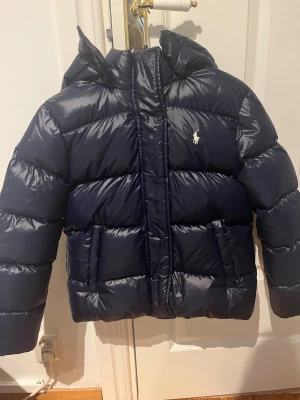 Mörkblå pufferjacka från Polo Ralph Lauren - Supernygg mörkblå pufferjacka från Polo Ralph Lauren. Jackan har dragkedja och tryckknappar framtill. Säljer den då den är för liten för mig och jag köpte den på plick men jag har aldrig använt den själv🥰Strl M/140 barn🙌🏻