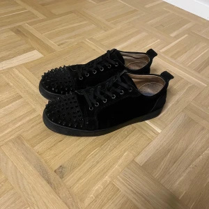Svarta Christian Louboutin sneakers med nitar - Svarta sneakers i mocka från Christian Louboutin med coola svarta nitar på tån och klassisk röd sula. (Röda sulan är inte tvättad) Skorna har snörning och rund tå, vilket ger en edgy vibe. Perfekta för dig som vill sticka ut med exklusiv design och detaljer. Storlek 43, hör av er vid minsta fundering!