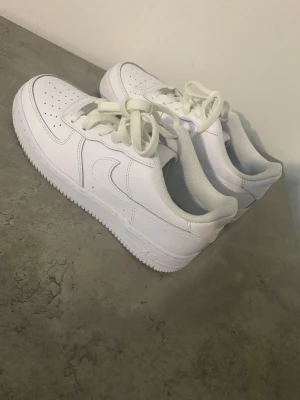 Nike Air Force 1 vita sneakers strl 38 - Fräscha Nike Air Force 1 sneakers i helvitt med klassisk design och låg profil. Skorna har snörning, perforerad tå och platt sula. Tillverkade i skinn med meshfoder för extra komfort. Perfekta för dig som gillar stilrena och ikoniska sneakers.