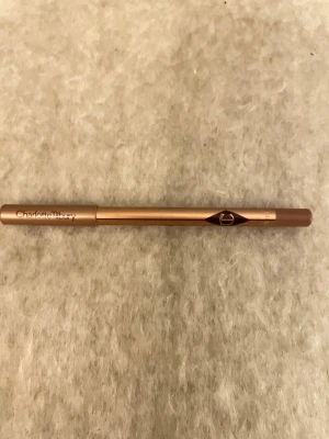 Charlotte Tilbury Iconic Nude läppenna - Säljer en Charlotte Tilbury Lip Cheat läppenna i färgen Iconic Nude. Pennan har en snygg roséguldig hylsa och en krämig nude-brun ton som är perfekt för att definiera och forma läpparna. Klassisk pennform i trä med lyxig finish.